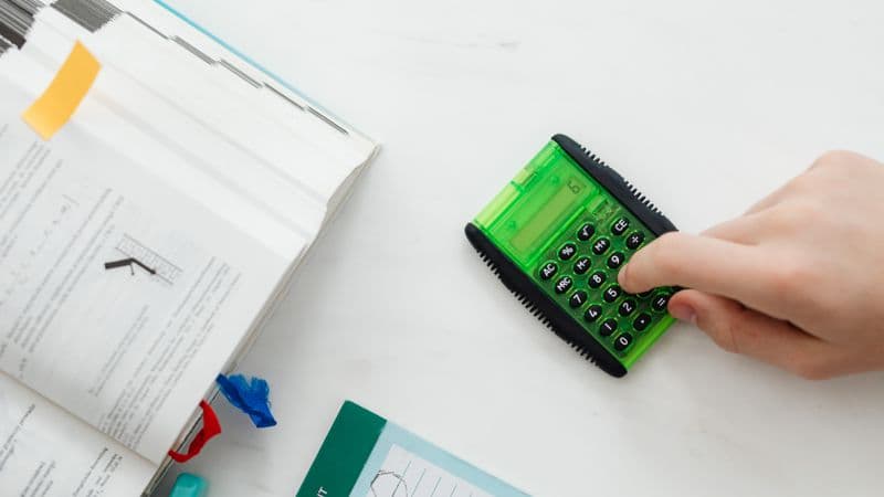 A student uses a green calculator to solve math equations from a textbook on a desk