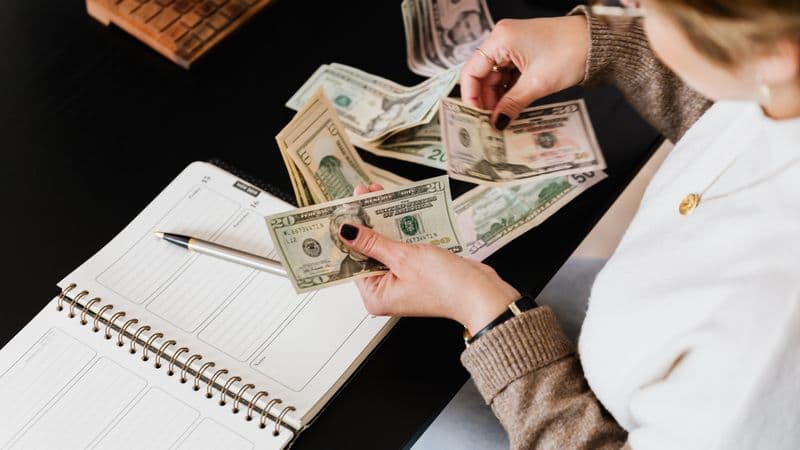 Woman organizing finances, counting dollar bills at desk with open notebook